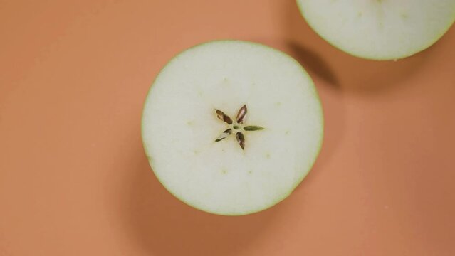 Top view shot of small green apple on isolated orange surface being cut in half with sharp steel knife. Upper fruit part falling beside. Food, healthy eating, cooking concept