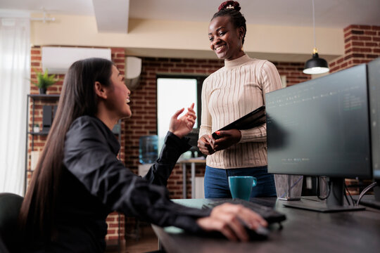 Happy smiling tech developing company workers laughing at funny hacker news story while sitting in modern workspace at computer desk. Multiracial coworkers enjoying break time together.