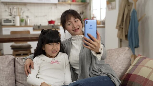 slow motion of happy asian mom and girl talking to friends through video conferencing at home. the woman holds mobile phone and sits close to her daughter on sofa