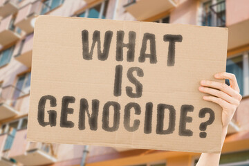 The question " What is genocide? " on a banner in men's hands with blurred background. City. Annihilation. Eradication. Street. Europe. Massacre. Destroy. Kill. Murder. Attack. Slaughter. Execute