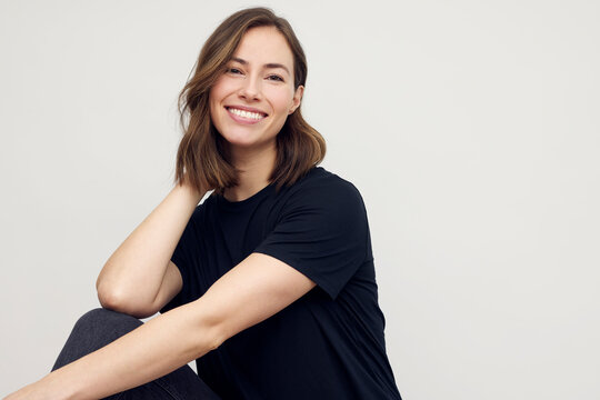 Portrait of a young happy woman smiling, looking beautiful and elegant. Looking in camera and sitting isolated on white background in a black t-shirt, touching her hair. 