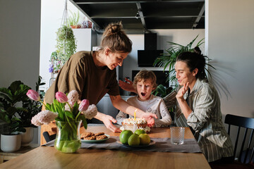 Happy lesbian family celebrating birthday of little son with birthday cake at home