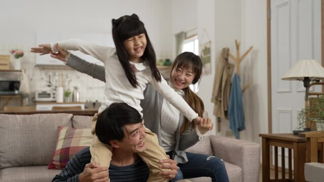 slow motion of excited Asian daughter playing and sitting on her dad’s shoulders in living room while her mom is holding her arms and having a good laugh