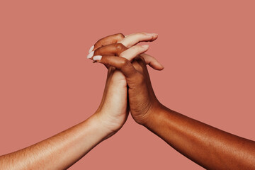 Close up multiracial woman couple with black and caucasian hands holding each other wrist in tolerance unity love and anti racism concept. Pink background.