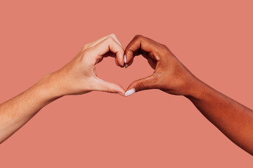 Close up portrait of black and white female hands in heart shape over pink background. Interracial...