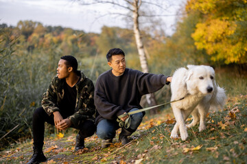 Portrait of asian and latin man sitting together and caressing Maremmano-Abruzzese Sheepdog while resting in nature at sunny autumn day. Concept of rest and weekend in nature