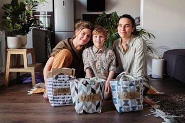 Portrait of homosexual mothers with little son looking at camera sitting on the floor with bags with sorted garbage for recycling