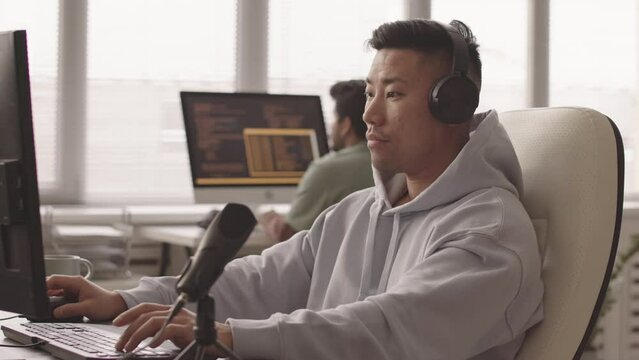 Waist up slowmo of Asian programmer in headphones sitting at desk with computer monitor and microphone in office developing video game on pc while his colleague typing program code in background