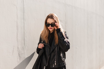 Fashion beauty girl hipster in leather black rock jacket and hoodie with bag wear stylish sunglasses and walks on the street near a gray wall