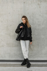 Stylish young woman model in trendy rock outfit with fashion leather jacket, jeans and black leather shoes with leather bag stands near a gray wall