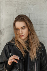 Fashionable urban portrait of young beautiful woman with hairstyle in black fashion leather rock jacket with bag stands near a gray concrete wall on the street