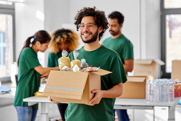 charity, donation and volunteering concept - happy smiling male volunteer with food in box and international group of people at distribution or refugee assistance center