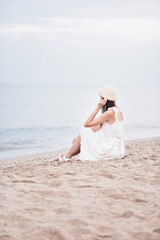 Fototapeta na wymiar Beautiful young girl sitting on the sand by the sea with her back turned in a white dress and hat