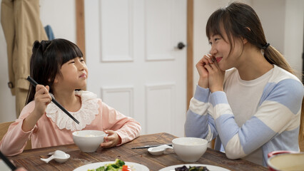 smiling asian mother looking her young daughter enjoying the meal she prepares at dining table at home. the girl saying yummy to her mom while eating