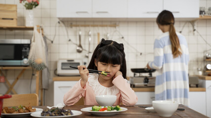 selective focus of unhappy picky Asian schoolgirl daughter holding face with disgust look on face while playing with food in bowl at dining table. her mother is cooking in kitchen at background
