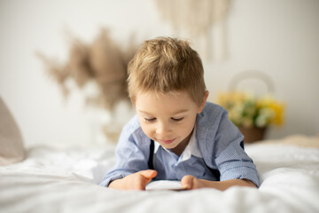 Blond preschool child, cute boy, playing on mobile phone in bed
