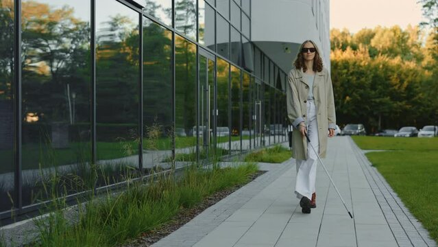 Beautiful young blind woman walking on city street with cane stick in hands. Caucasian lady in casual clothes checking safety direction outdoors.