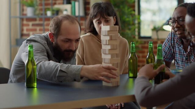Positive people having fun with jenga board game in living room, playing with wooden tower blocks. Men and women enjoying game with building square pieces on structure for entertainment.