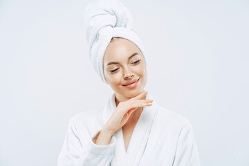 Pretty natural woman touches gently jawline, wears bath towel wrapped on head, dries hair after shower, cares about body, has clean skin, prepares for evening party