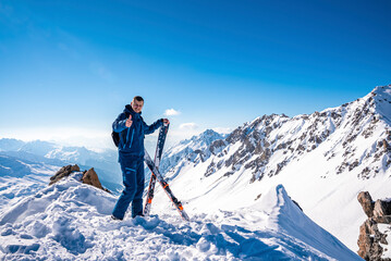 Fototapeta na wymiar St. Anton am Arlberg. March 10, 2022. Young man showing thumbs up while holding skis on top of snowy mountain, Skier show thumbs up on the background of beautiful view of snowcapped mountains