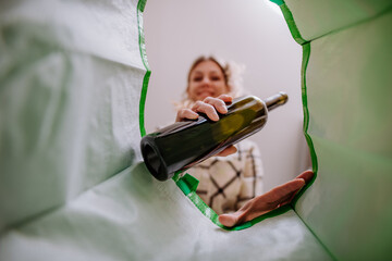 Image from inside green recycling bag of woman throwing a glass bottle to recycle.