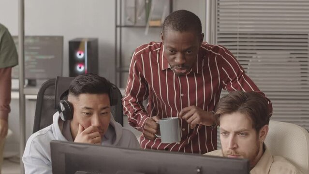 Medium slowmo of diverse team of programmers looking at computer monitor and talking while developing new software or video game, coding and programming together in modern office