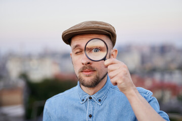 Male searching sales or discounts at sunset using loupe with urban view. Shopping or consumerism concept. Cheerful man looking with magnifying glass at camera wearing jeans shirt and peaked cap