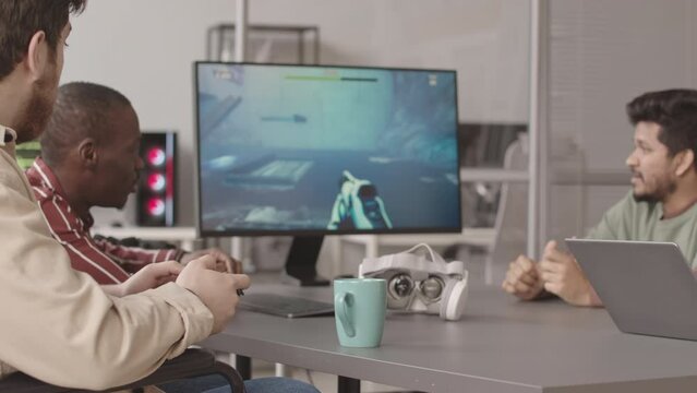 Medium slowmo of diverse team of programmers looking at computer monitor while testing design of first person shooter game they developed together