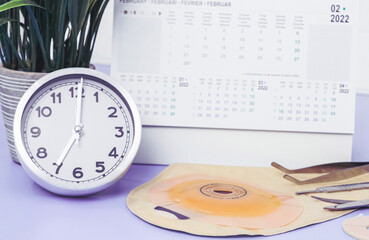 Clock with time seven, calendar for February 2022, surgical instruments, colostomy bag on a lilac.