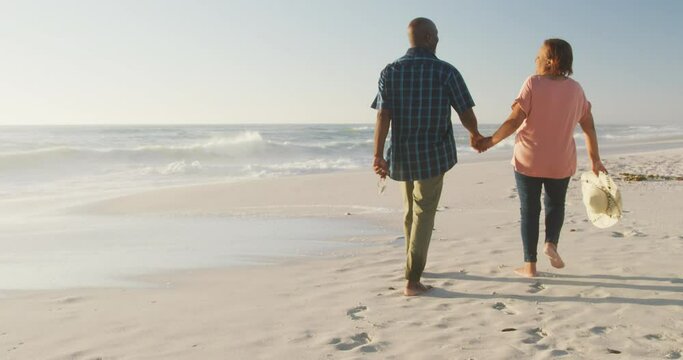 Smiling senior african american couple holding hands and walking on sunny beach