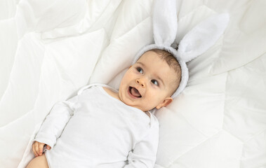 super cute little baby with white bunny ears on head laying on white blanket. toddler is smiling,...