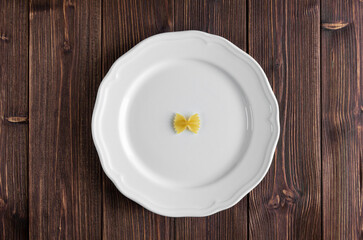 One raw pasta farfalle on white plate on wooden table.