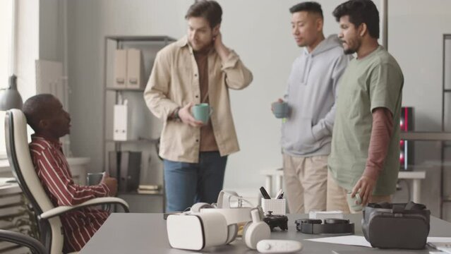 Medium slowmo of two vr headsets on table in foreground and multiethnic team of vr games developers talking during coffee break, working together in modern office