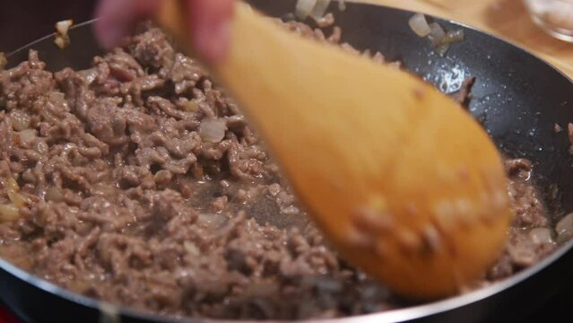 Close up shot of female hand using wooden spoon during browning ground beef meat in hot skillet