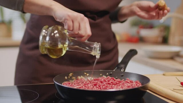 Close up shot of woman in apron pouring olive oil over ground beef meat in frying pan while preparing food in kitchen