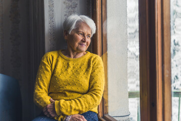 Sad Caucasian Grey Haired Senior Pensioner Looking through the window with winter scenery - Smiling but Sad eyes. High quality photo