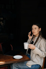 A young European freelancer girl with long dark hair in a shirt remotely works in a cafe, businesswoman are talking to colleagues on the phone, drinking black coffee, and looking on the street