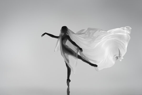Black and white portrait of graceful ballerina dancing with fabric, cloth isolated on grey studio background. Grace, art, beauty concept. Weightless, flexible.