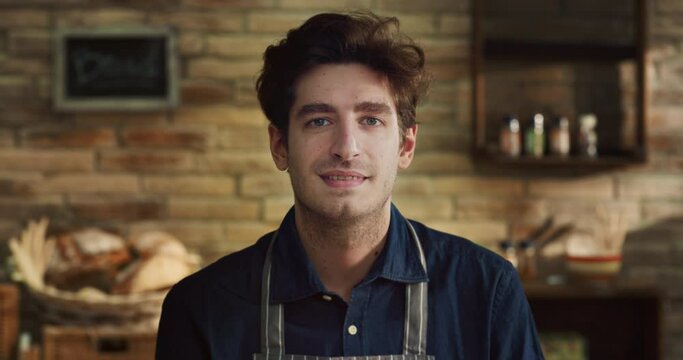 Cinematic close up shot of young male artisan baker is smiling in camera while kneading dough for preparation of pasta, pizza and other pastries in rustic bakery shop kitchen.
