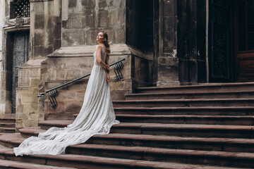 Beautiful bride in a long white wedding dress stands on the stairs near the old church