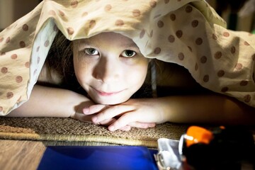 Cute little girl hides under a blanket and reads a fairy tale about a magical space world