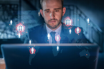 Handsome businessman in suit at workplace working with laptop to hire new employees for international business consulting. HR, social media hologram icons over office background