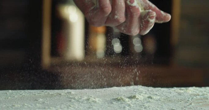 Cinematic macro shot of professional artisan baker chef sprinkles flour dust powder on table while kneading dough for preparation of pasta, pizza and other pastries in rustic bakery kitchen.