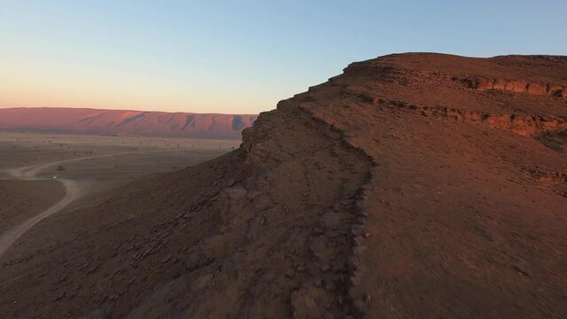 Drone flying over rocky formation in desert with red mountain in background at sunset, Morocco. Aerial fpv
