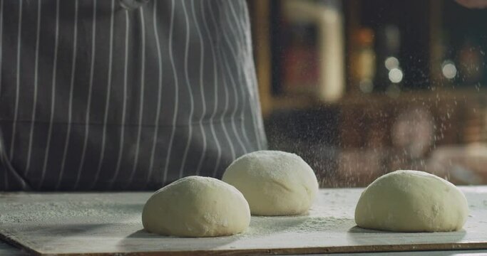 Cinematic close up of professional artisan baker chef sprinkles flour dust powder on fresh just prepared loaves of dough for preparation of pasta, pizza and other pastries in rustic bakery kitchen.