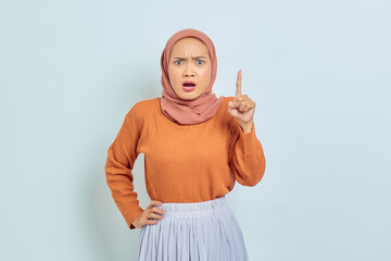 Naklejka premium Surprised young Asian Muslim woman in brown sweater pointing to copy space with finger isolated over white background. Muslim lifestyle concept