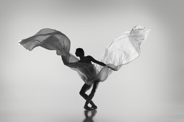 Black and white portrait of graceful ballerina dancing with fabric, cloth isolated on grey studio background. Grace, art, beauty concept. Weightless, flexible.