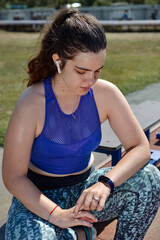 Outdoors on a sunny hot day, a young sweaty woman in wet clothes goes in for sports, takes a break, counts the pulse using an electronic watch, listens to music in wireless headphones.