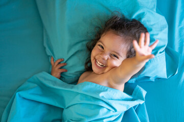 Obraz na płótnie Canvas Niña hermosa juguetona sonriente y feliz acostada descansando y disfrutando y jugando recostada en la cama con sábanas color azul y cobijada