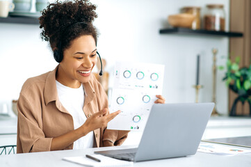 Successful smart african american curly-haired young woman, in casual wear, working or studying from home, using laptop and headset, talking on video conference, conducts financial consultation, smile